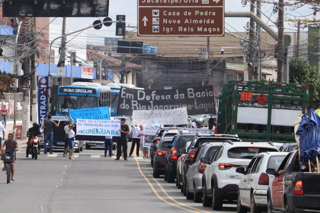 Thumbnail:  Esta é uma foto de uma cena urbana diurna, tirada ao nível da rua, que captura um protesto bloqueando o tráfego em uma avenida movimentada.

              No centro da imagem, um grupo de manifestantes está de pé no meio da rua, segurando faixas e cartazes. A faixa principal, grande e preta com letras brancas, atravessa a largura da via e diz: 'Em Defesa da Bacia do Rio Jacaraipe-Lagoa'. Há também cartazes menores, incluindo um azul claro que diz 'NÃO AO ATERRO/CALHAMENTO E INUNDAÇÃO DOS RIOS'.
              
              Devido ao protesto, o trânsito está parado. No lado direito da foto, há uma longa fila de carros enfileirados. O carro mais próximo em primeiro plano é um SUV branco, seguido por outros veículos de passeio e, mais atrás, um caminhão verde com carroceria de ripas.
              
              No lado esquerdo da rua, um ônibus azul de transporte público, com a identificação '806A CIRCULAR' em seu letreiro digital, está parado atrás dos manifestantes. Algumas motocicletas também estão paradas nas proximidades. Na extrema esquerda, em primeiro plano, uma pessoa anda de bicicleta pela rua.
              
              Acima de toda a cena, paira uma complexa rede de fios e cabos elétricos. Há também uma grande placa de trânsito marrom suspensa sobre a rua, com setas e nomes de locais como 'Jacaraípe/Orla', 'Casa de Pedra' e 'Nova Almeida'. Ao fundo, podem-se ver edifícios e a continuação da paisagem urbana. O céu está claro, mas nublado, indicando um dia sem sol forte.
              
              A atmosfera geral é de uma manifestação civil, causando uma interrupção na rotina da cidade para chamar a atenção para uma causa ambiental.; disponível em https://www.instagram.com/p/DNfuk5htYXX/?img_index=1