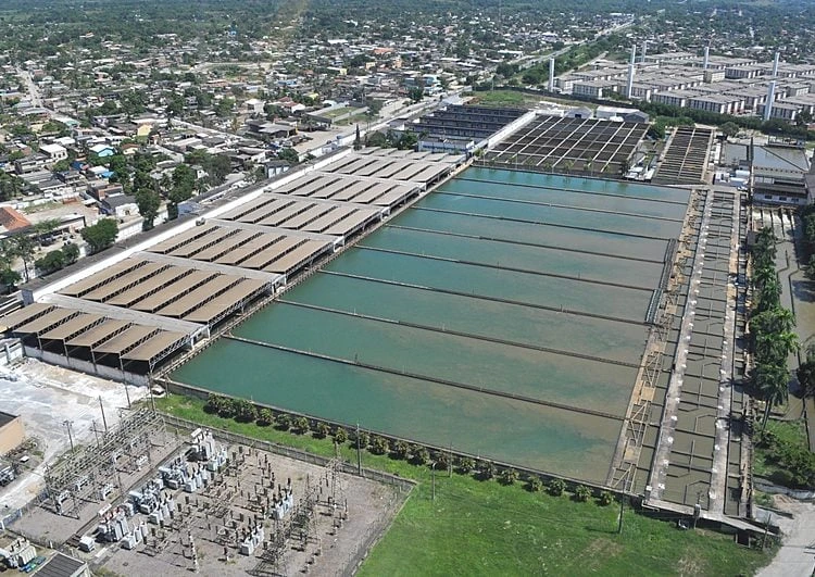 Thumbnail: Esta é uma imagem aérea de uma ampla e complexa instalação industrial, que se trata de uma Estação de Tratamento de Água (ETA) ou de Esgoto (ETE), uma peça fundamental para o saneamento básico e o abastecimento de uma cidade.
O ponto focal da fotografia são os grandes tanques retangulares que dominam o centro da imagem. Eles estão organizados em fileiras paralelas e contêm água em diferentes estágios do processo de purificação. A coloração da água varia de um tanque para o outro, indo de tons mais turvos e esverdeados a outros mais claros, indicando fases como floculação, decantação e filtração. É possível ver passarelas e divisórias entre os tanques, utilizadas para operação e manutenção. À esquerda, uma série de tanques menores está coberta por estruturas, sugerindo uma etapa diferente do tratamento.
No primeiro plano, à esquerda, destaca-se uma subestação de energia elétrica, com seus transformadores, isoladores e toda a fiação necessária para alimentar a operação de alta demanda da estação de tratamento.
Ao fundo e ao redor da instalação, estende-se uma vasta área urbana, com centenas de casas e edifícios, evidenciando a população que depende diretamente desta infraestrutura para ter acesso à água potável e saneamento. A imagem ilustra de forma clara a interface entre a engenharia sanitária, os recursos hídricos e a vida urbana, ressaltando a importância vital do tratamento de água para a saúde pública e a qualidade de vida.