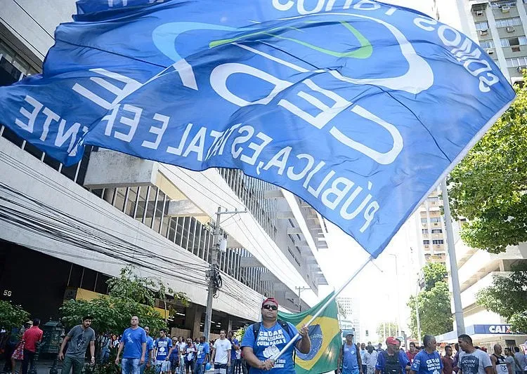 Thumbnail:  Esta imagem retrata uma manifestação em uma rua urbana, onde um grupo de pessoas, provavelmente trabalhadores, protesta contra a privatização da CEDAE. Em destaque, um homem carrega uma grande bandeira azul com os dizeres CEDAE PÚBLICA, simbolizando a luta pela manutenção da empresa como estatal, enquanto outros manifestantes o seguem, reforçando a mensagem coletiva do ato; disponível em https://assets.brasildefato.com.br/2024/09/image_processing20200201-29235-15ef41c.jpg